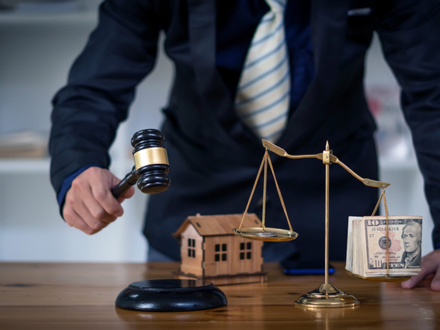 A person in a dark suit holds a judge's gavel in their right hand, positioned above a sounding block on a wooden table. To their side, there's a small model of a house and a balance scale with a stack of U.S. currency on one side.