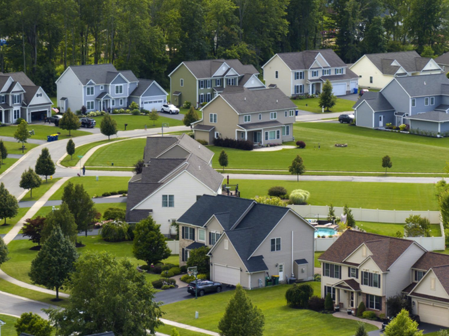 An aerial view of a suburban neighborhood shows a collection of two-story houses, each with its own well-manicured lawn. The homes are architecturally similar, with variations in color schemes and details, presenting a cohesive yet individualized community aesthetic.