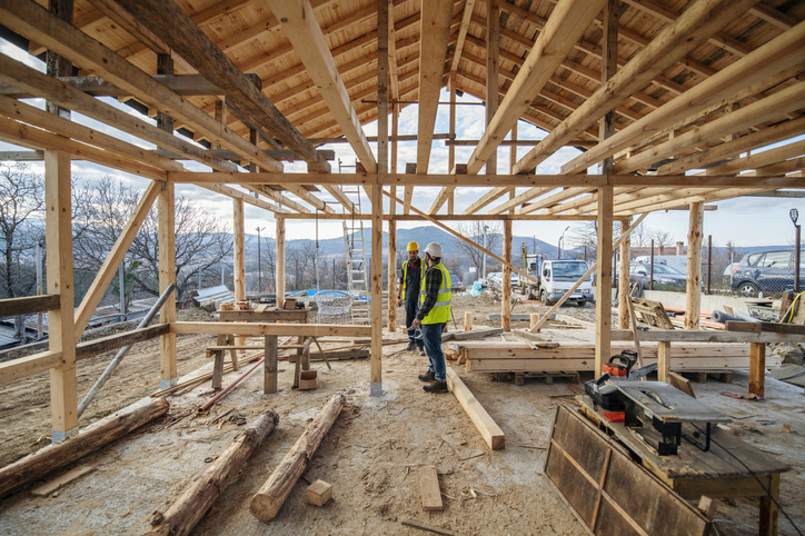 Two construction workers in high visibility jackets are discussing plans inside the wooden frame of a house under construction, surrounded by tools and building materials.