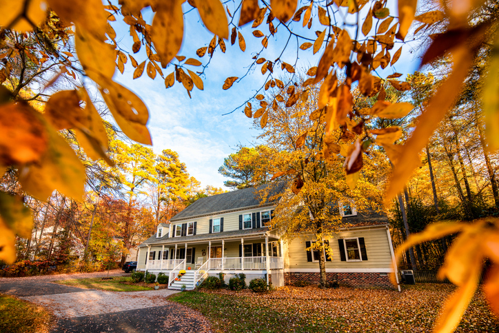 A picturesque two-story house is framed by the vibrant foliage of autumn. The home's classic design, complete with a covered front porch and dormer windows, is nestled amidst a landscape covered with a carpet of fallen leaves.