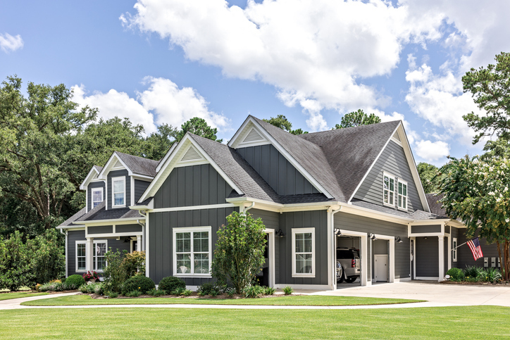 A modern gray house with white trim and an attached garage, set against a backdrop of lush trees and a well-maintained lawn, complete with an American flag displayed prominently.