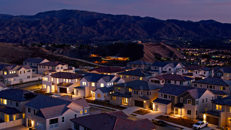 A residential area with modern homes illuminated by evening lights, nestled against a backdrop of rolling hills under a twilight sky.