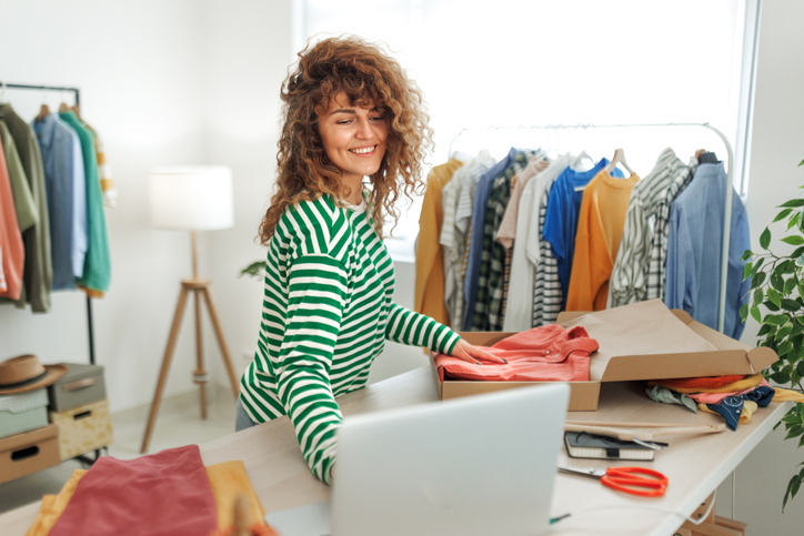 A cheerful woman with curly hair, wearing a striped shirt, is packaging clothes in a box, with a laptop open in front of her.