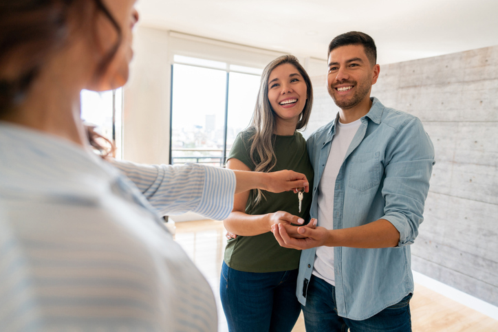 A happy couple is receiving keys from a real estate agent, as they stand in a bright, modern apartment.