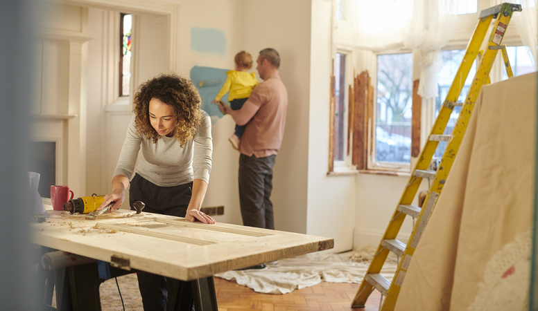 A woman is focused on a DIY woodworking project on a sturdy table in a room that appears to be undergoing renovations. In the background, a man is holding a baby, standing next to a yellow ladder and painting the wall with a roller.