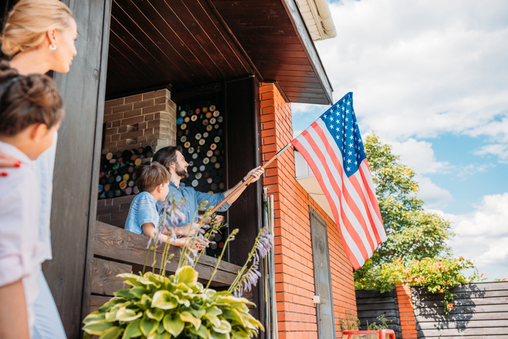 A family is engaged in a patriotic moment, with a man and child holding the American flag out from the porch of their home, as others watch with pride.