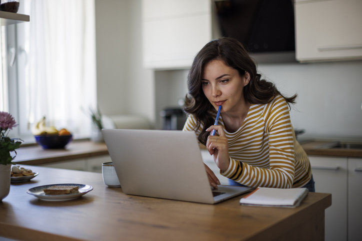 A pensive woman in a striped shirt works on her laptop at a kitchen table with a notepad and a pen in her mouth, with a meal set beside her.